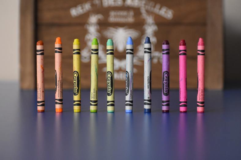 A row of crayons in rainbow colors stands on end, with a blurred wooden crate in the background.