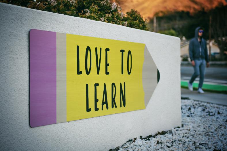 The picture shows a student in jeans and a hoodie, walking past a sign depicting a yellow pencil with a purple eraser inscribed with the words, "Love to Learn."