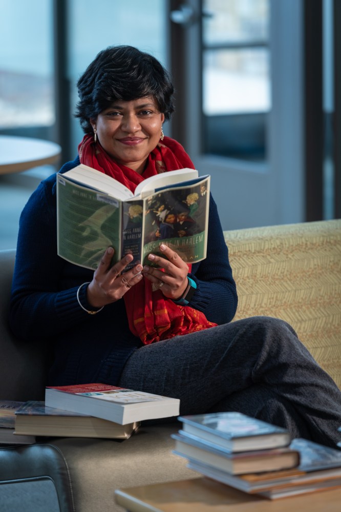 Library employee sits on a sofa with an open book in her hands, smiling at the camera. Other books are piles around her.