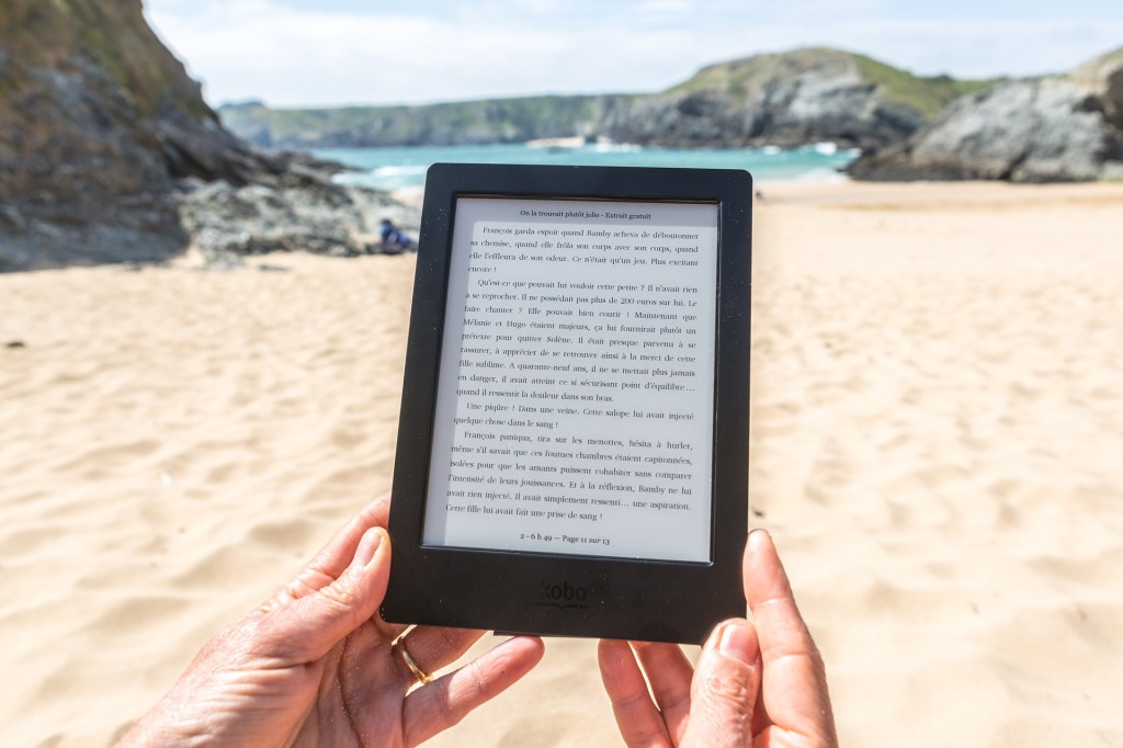 The image shows a pair of hands holding an ereader with a remote sandy beach, rocky hills, and a turquoise sea and hazy blue sky in the background.