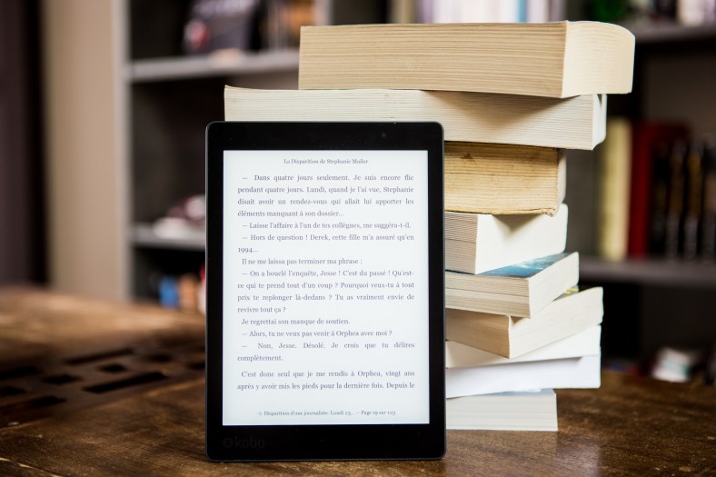 The image depicts an ereader on a wooden table, leaning up against a stack of nine paperback books, with a bookcase in the background.