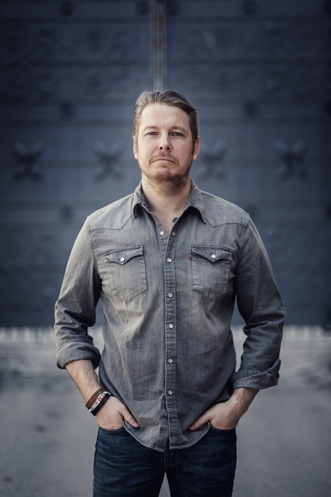 The author, dressed in a dark grey button down shirt, stands with his hands in his jeans pockets. He has short brown hair, and a slight beard.