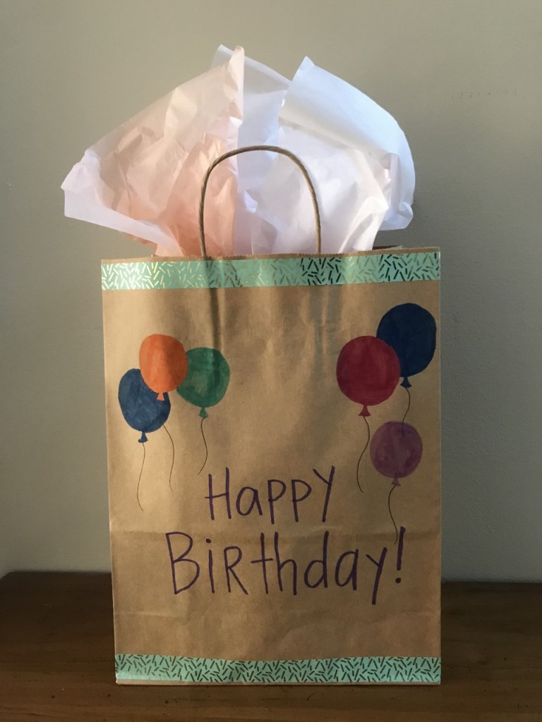 Brown paper bag decorated with marker and tape to say Happy Birthday, with tissue paper sticking out from the top.