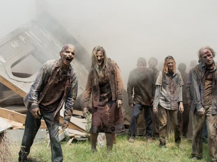 A crowd of zombies stagger past the wreck of a building, coming out of dense fog.