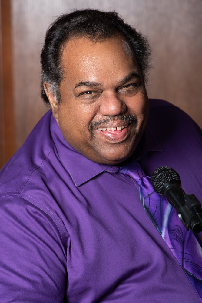 Headshot of a smiling Daryl Davis, wearing a purple shirt and tie, seated in front of a microphone.