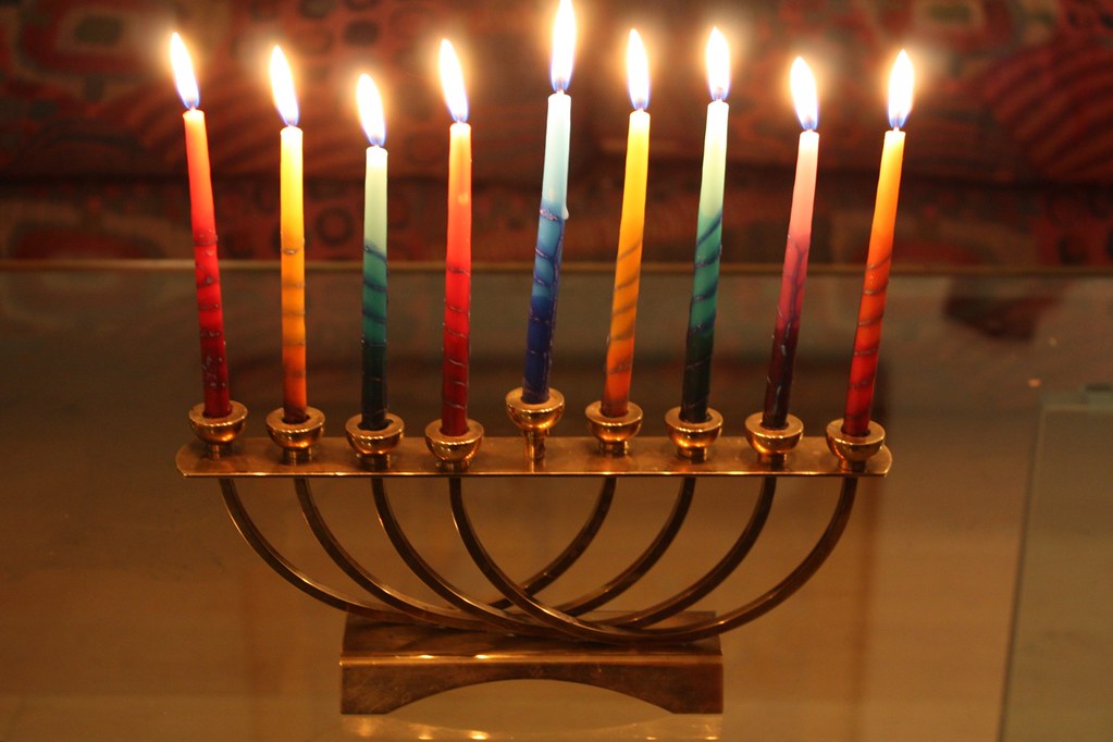The photograph depicts a tabletop menorah with nine lit candles in primary colors.