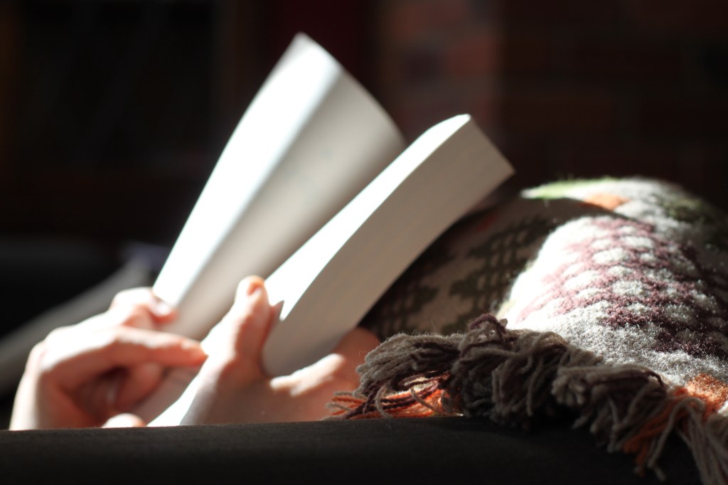 The photograph depicts two hands holding an open book against a colorful woolen blanket, with sunlight streaming in to illuminate the book.