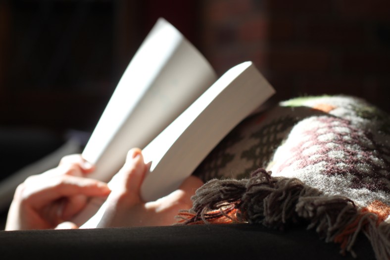 The photograph depicts two hands holding an open book against a colorful woolen blanket, with sunlight streaming in to illuminate the book.