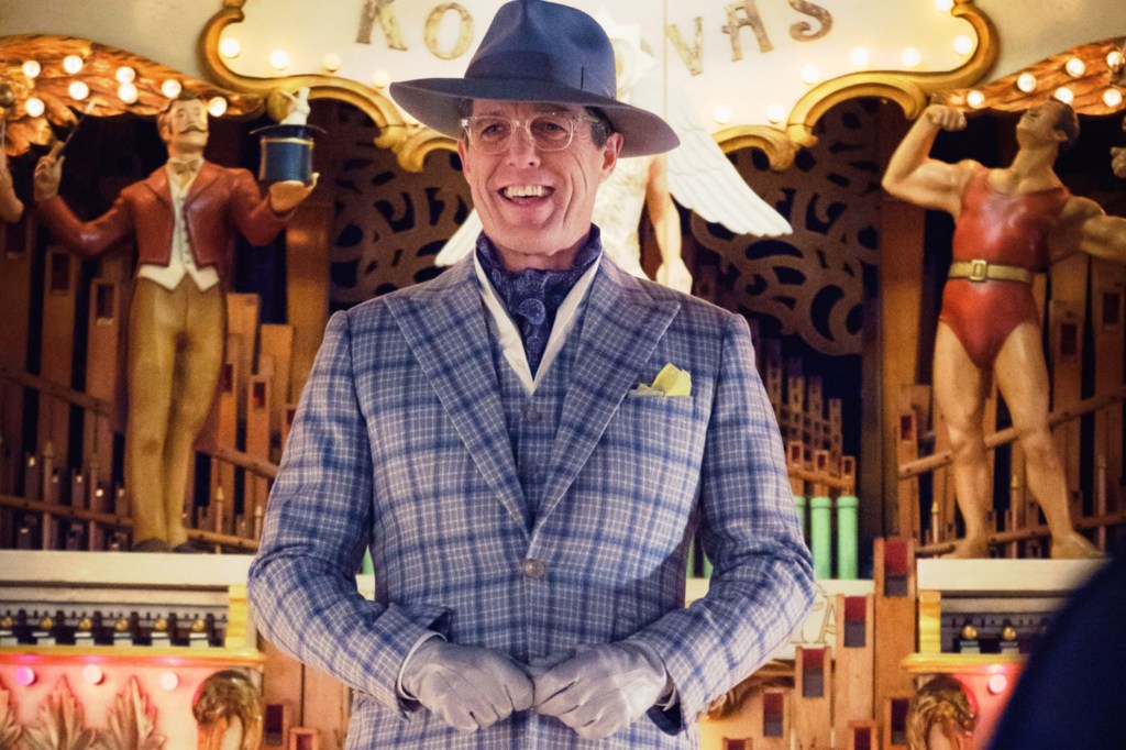 Hugh Grant is depicted in a blue and grey checked suit with ascot, and a blue wide-brimmed fedora, in front of a carnival-themed calliope.