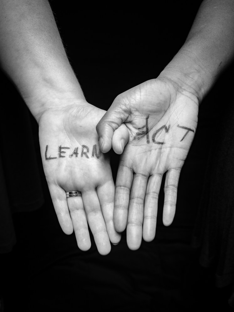 Two hands joined by hooking thumbs. The hand on the left is White and has the word "Learn" on the palm. The hand on the right is Black and says "Act."