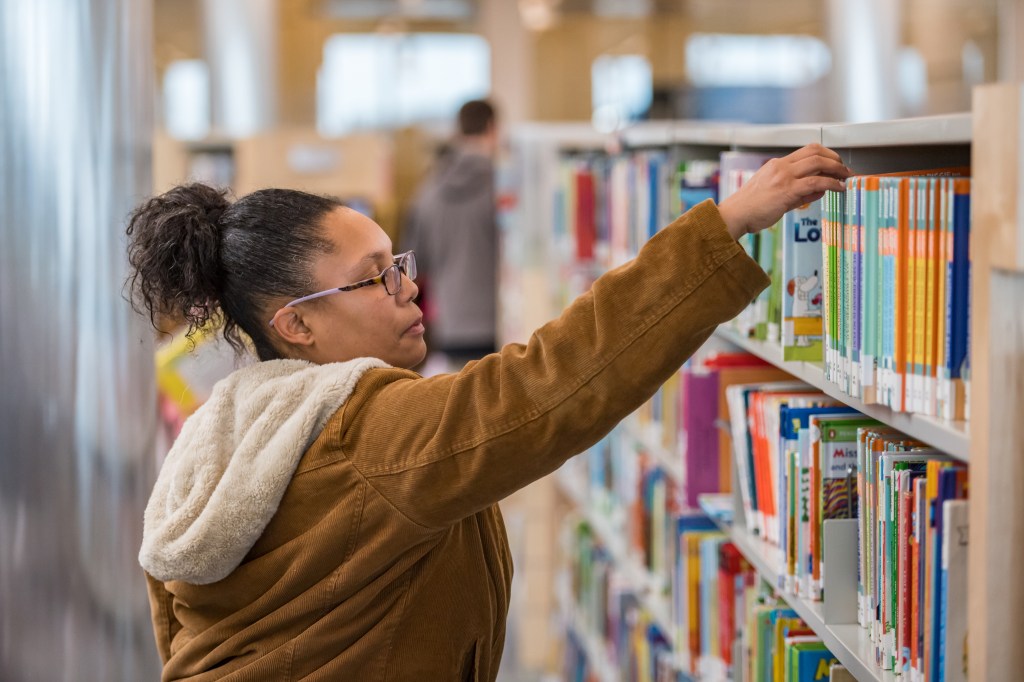 A woman, with a ponytail and glasses and wearing a light brown corduroy coat, reaches for a book off a library shelf.