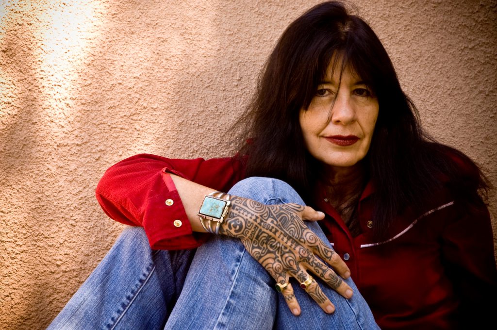 The photograph depicts poet laureate Joy Harjo wearing a bright red shirt and blue jeans, with her tattooed hand across her knee and a turquoise bracelet on her wrist.