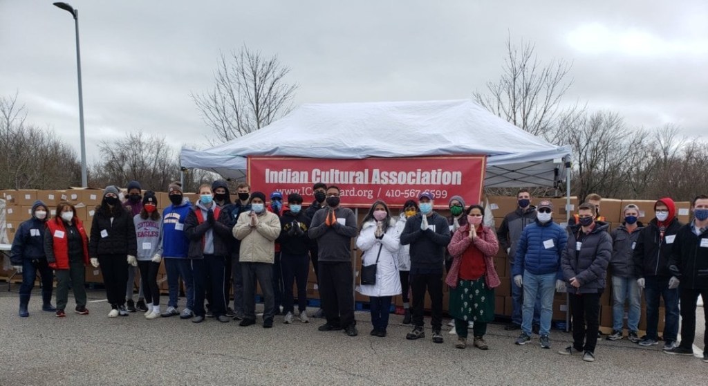 A group of people in winter coats and masks stand in front of a white pop-up tent, from which hangs a red banner that read Indian Cultural Association.