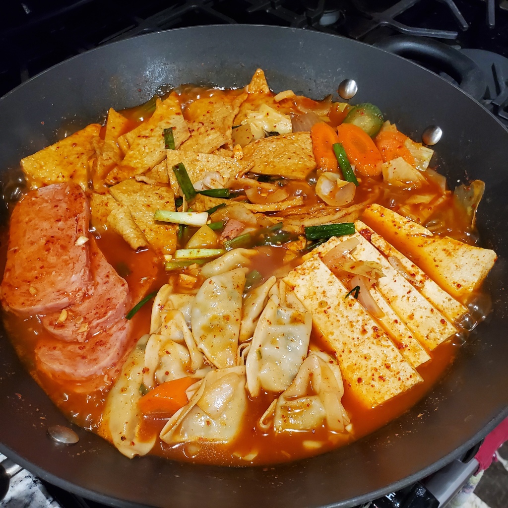 The photograph of budae jjiajae shows a pan of kimchi-based stew with spam, vegetarian dumplings, tofu, and fish cakes in a spicy red pepper sauce.