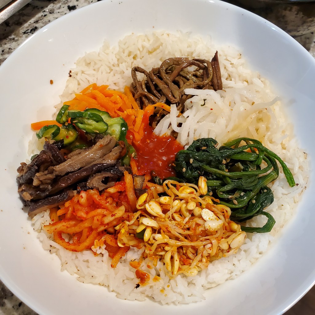 The photograph is of bibimbap on a white plate containing bean sprouts, spinach, pickled carrots, cucumbers, pickled radish, mushrooms, and red pepper sauce on a bed of white rice.