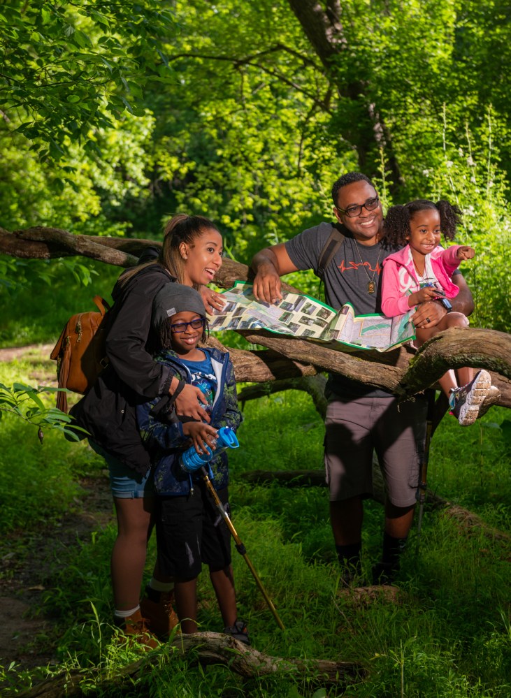 A family stands in a local park with a map spread out along a low horizontal tree branch.