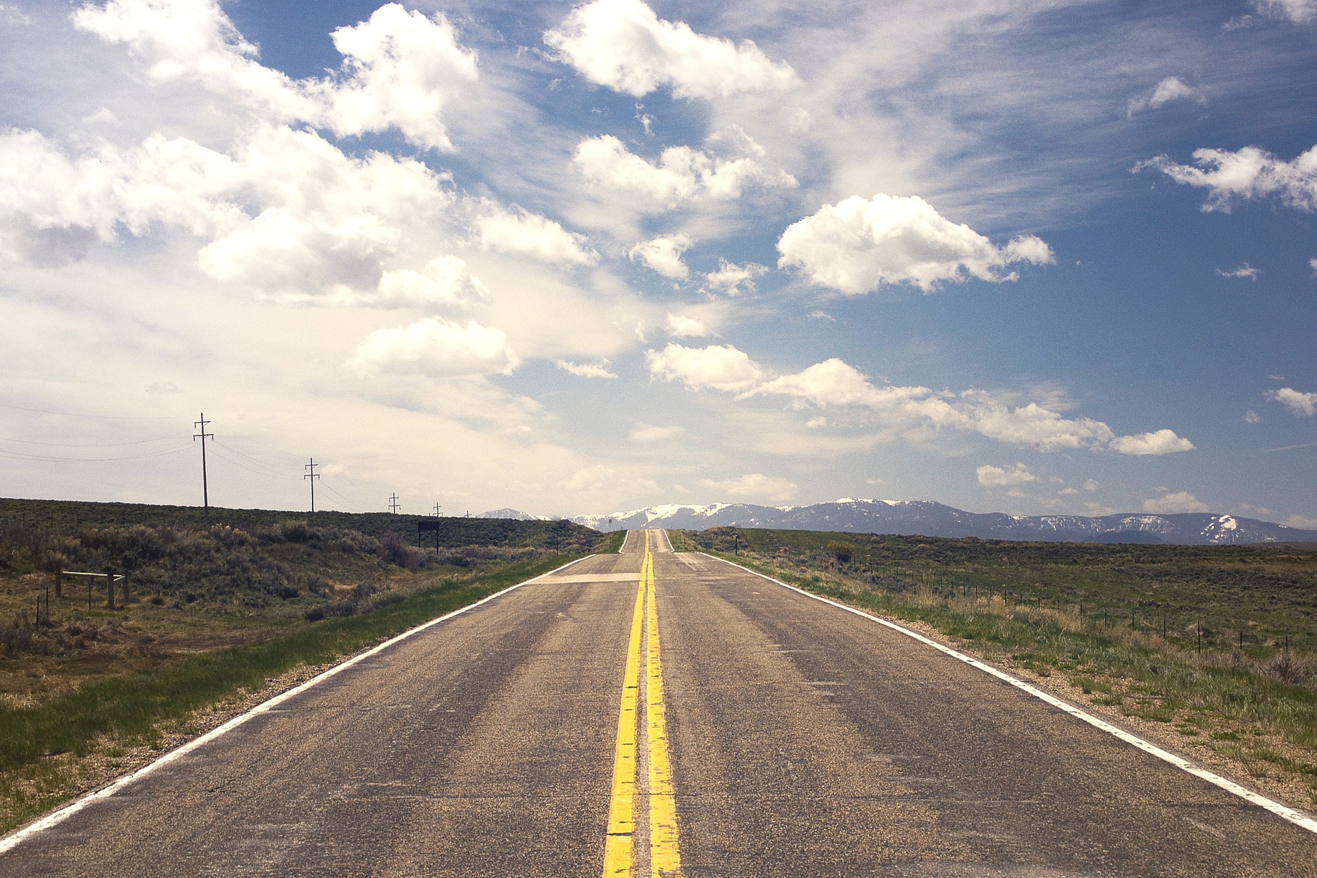 Open two-lane road with double yellow line leading into the distance of snow-topped mountains. Bright sky with white clouds take up the top two thirds of photo.