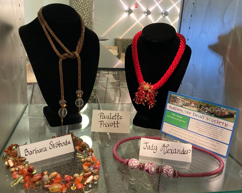 A picture of the Baltimore Bead Society collection in the Miller Branch display cases, with an assortment of beaded necklaces in red, pink, earth tones, and gold (clockwise from upper right corner).