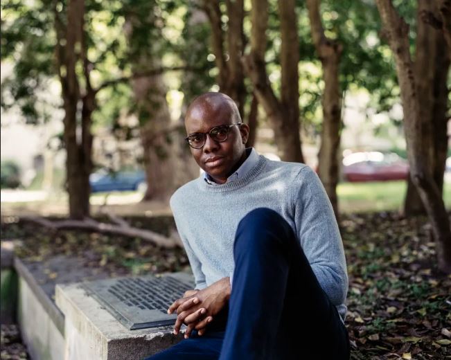 Young Black man dressed in dark blue pants and a grey sweater show seated in a park, leaning against a memorial black. He is bald and wears glasses.