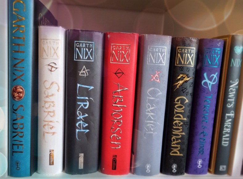 A shelf of books all in same style showing a majority of Garth Nix's Old Kingdom series: Sabriel in blue, Sabriel in white, Lirael in black, Abhorsen in red, Clariel in grey, Goldenhand in black, Terciel & Elinor in purple.