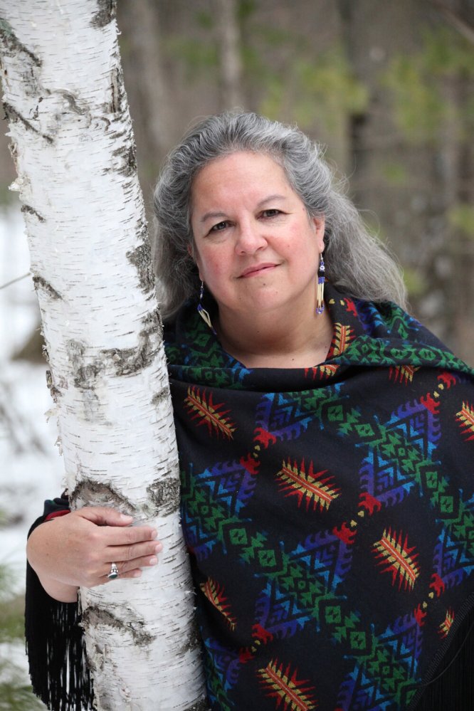 The author, with long grey hair pulled away from her face, leans against a white birch tree. She is wearing a richly colored and patterned poncho and dangling beaded earrings.