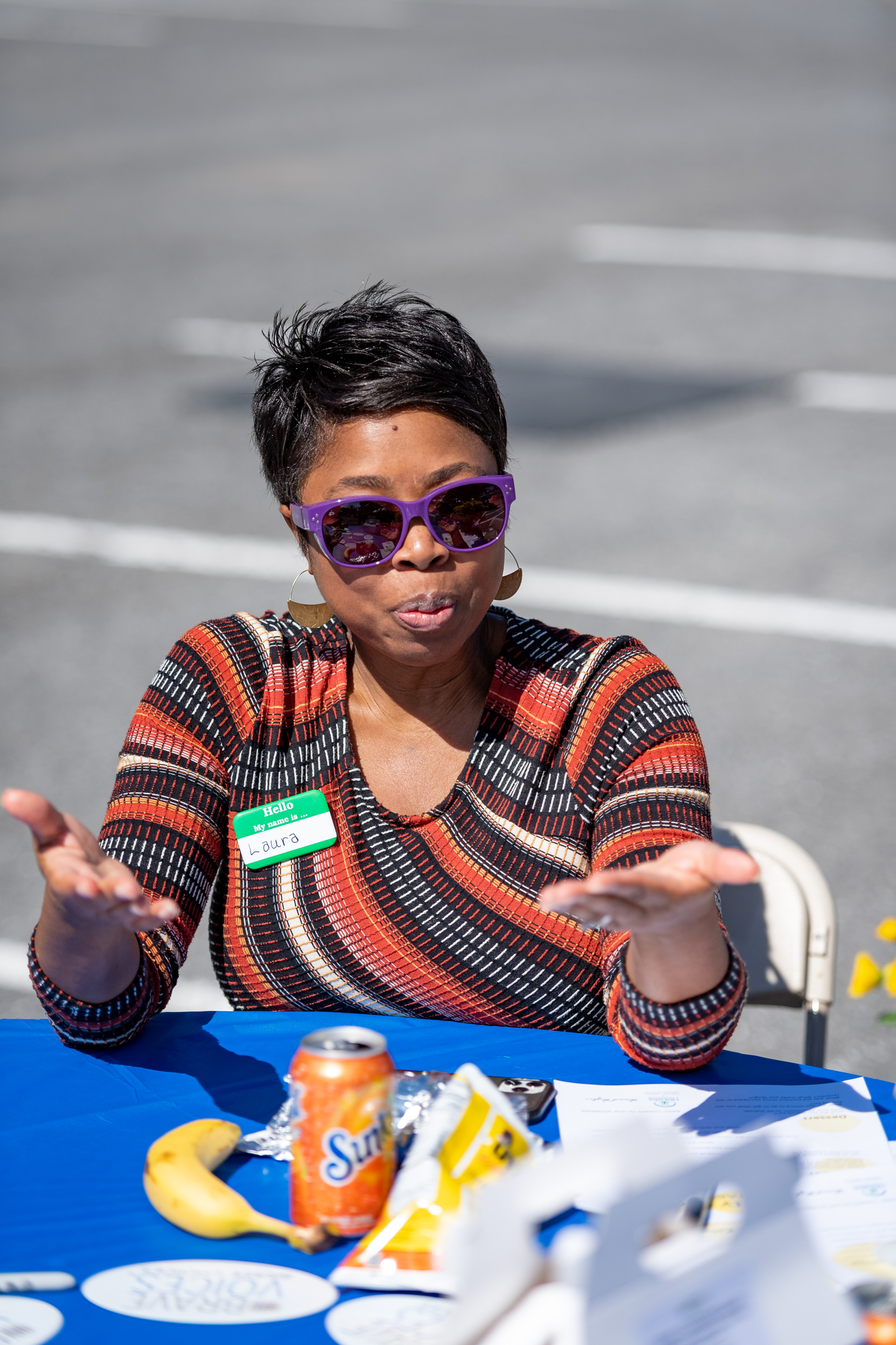 A Black woman with short hair, wearing purple sunglasses, gestures while sitting at a table.
