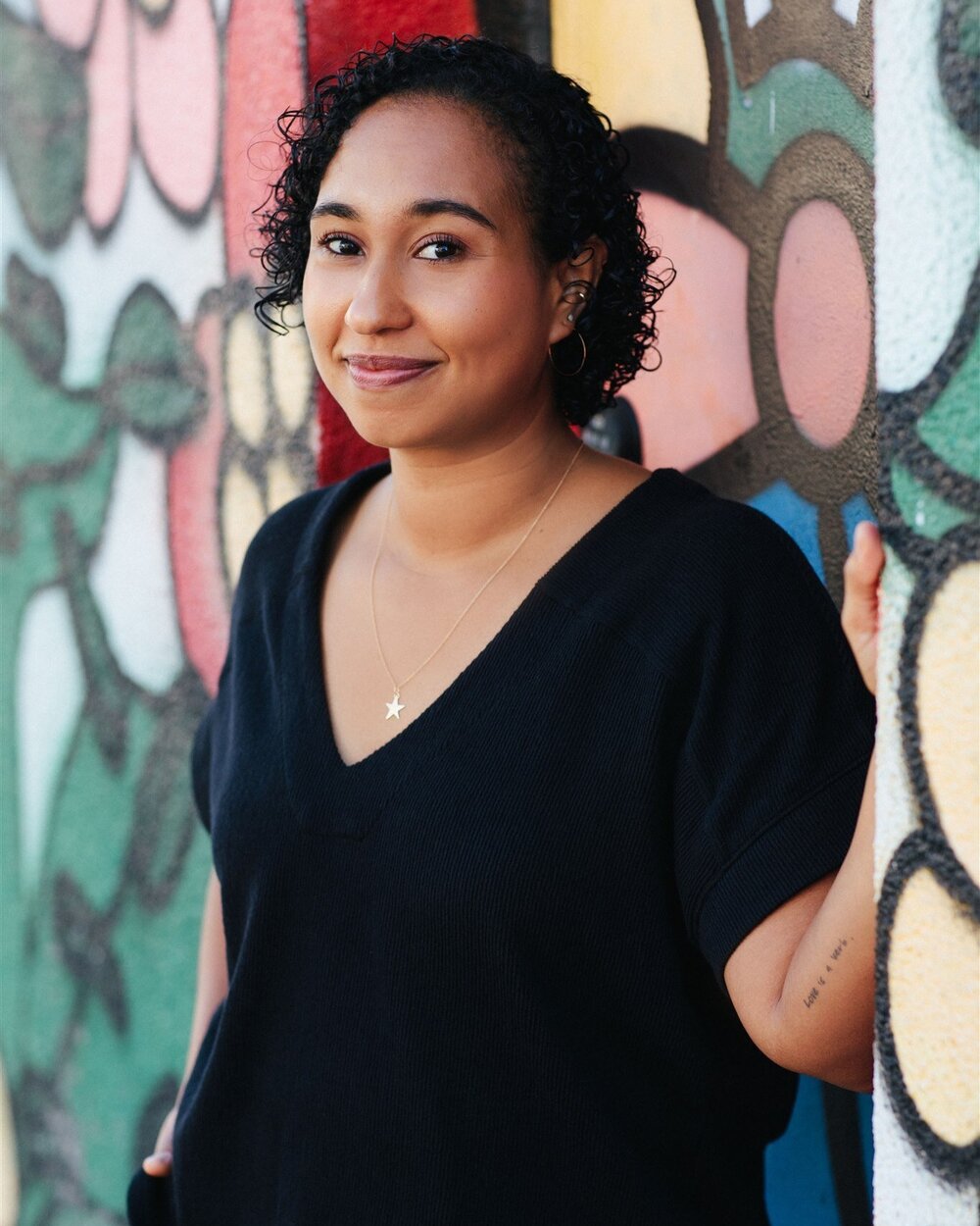 A young Black woman with short curly hair, wearing a black V-neck shirt stands by a wall painted in flowers.