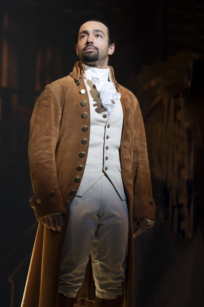 The photograph depicts actor Pierre Jean Gonzalez in his starring role as Alexander Hamilton, wearing a white shirt, pants, vest, and cravat, with a brown overcoat with gold buttons. His head is turned to the side and he is gazing into the distance.