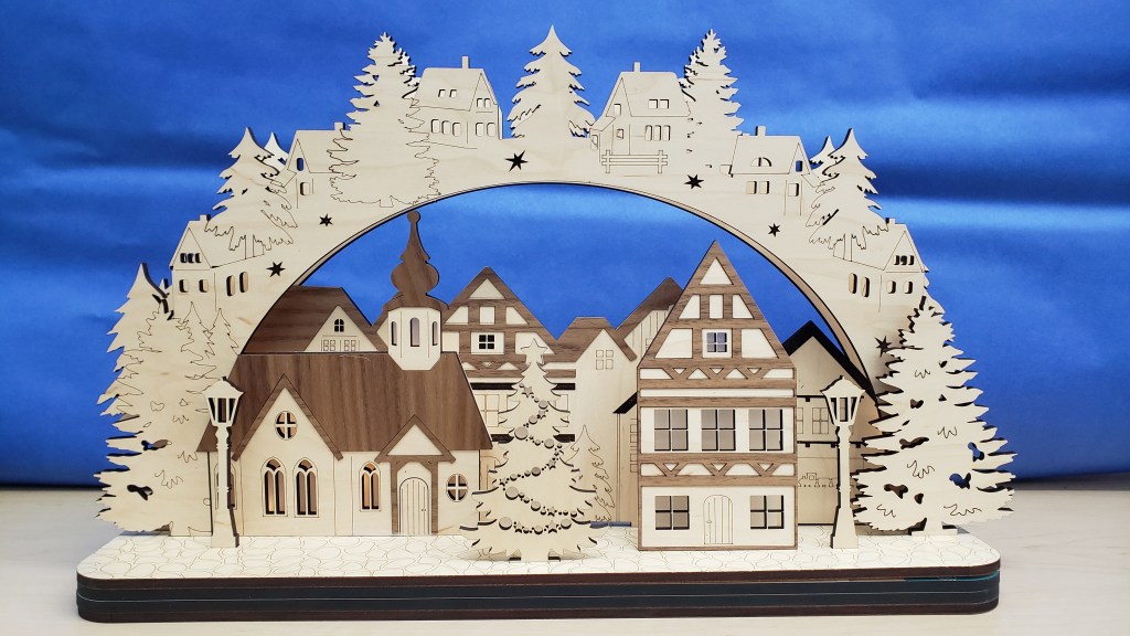 A picture of a German Schwibbogen village scene made with the laser cutter. The photograph consists of a village with homes, a church, lampposts, and fir trees, surrounded by an arch that has more cottages and fir trees, against a blue background resembling the sky.