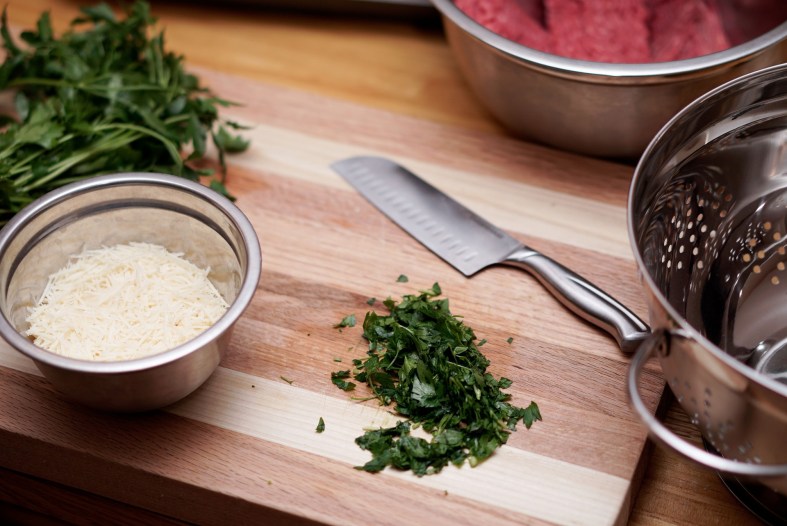 A wooden cutting board holds chopped green herbs, a stainless steel knife, and a small metal bowl with grated cheese. Nested colanders sit next to the board.