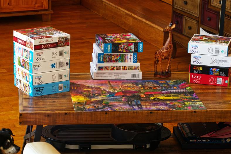 The photograph shows a wooden table with an assortment of 500- and 1000-piece puzzles in boxes on top. A puzzle with a rainy street view of Paris, with the Eiffel Tower in the background, is put together on the table surface. A wooden giraffe also stands on the table.