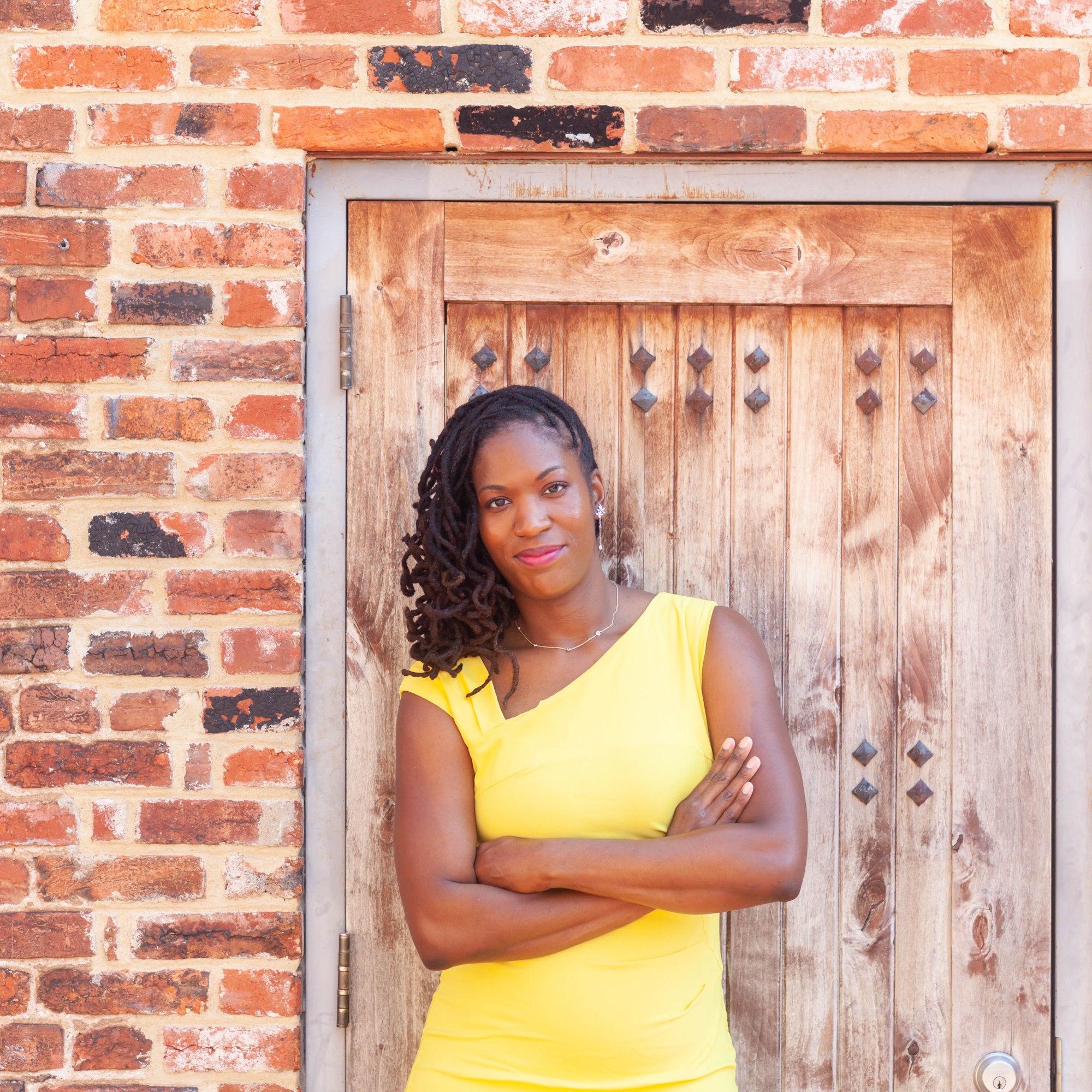 Faye McCray, wearing a yellow dress, stands before a wood door in a brick wall. She has her arms crossed and her hair all to the left.