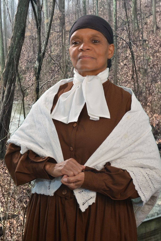 Storyteller and keynote speaker Janice Curtis Greene in costume as Harriet Tubman, wearing a brown dress, and white lace shawl, a white jabot, and a black headwrap.