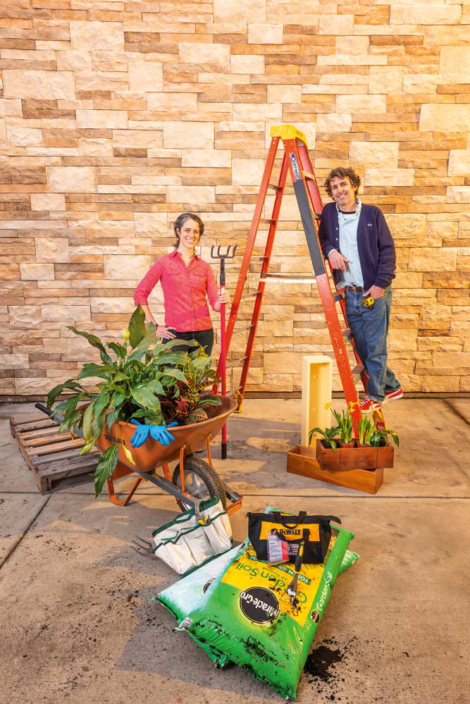 Eric stands on the bottom rung of a ladder, while Kelly holds a garden rake, with a stone wall behind them. A wheelbarrow full of greenery, a box of daffodils, and other gardening implements rest in front.