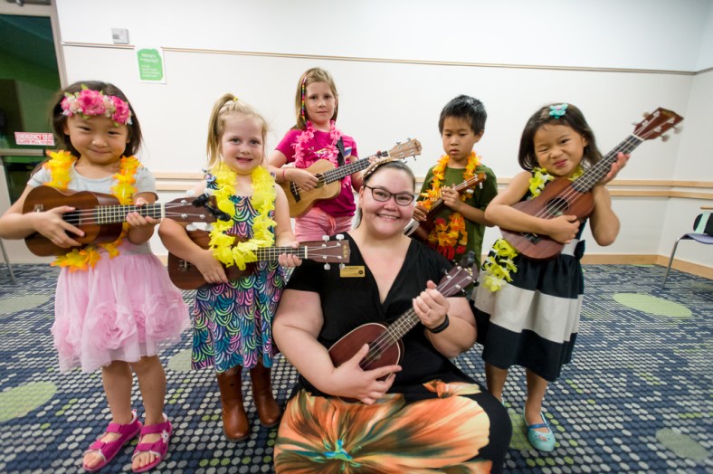 Several children stand around a library instructor, everyone strumming a ukulele.