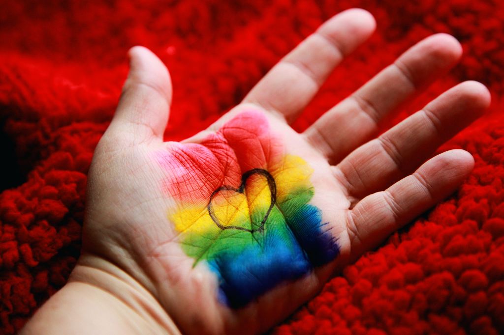 The photograph depicts a hand outstretched against a background of fluffy, bright red material. A rainbow of primary colors is painted on the hand, with the outline of a heart overlaid in black.