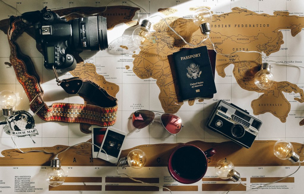 The photograph shows a map of the world with various objects lying across it, including cameras, a passport, photographs, sunglasses, a string of light bulbs, and a coffee cup.