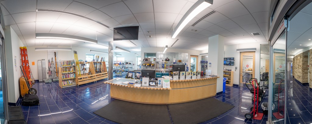 The photograph centers the customer service desk at the Elkridge branch's makerspace, with a wheelbarrow, ladders, and an array of books and tools to the left and behind, and a dolly and other equipment to the right.