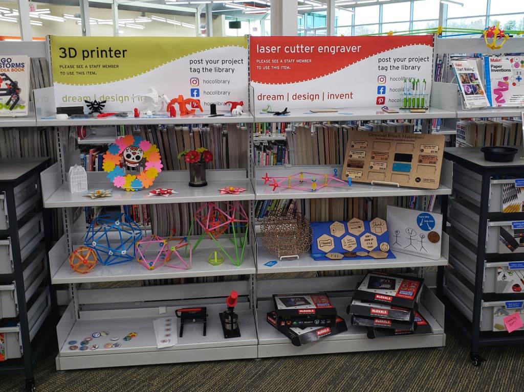 Signs advertising a 3D printer and laser cutter engraver, with examples of projects created by them on the shelves beneath, including keepsake ornaments and 3D printed flowers.