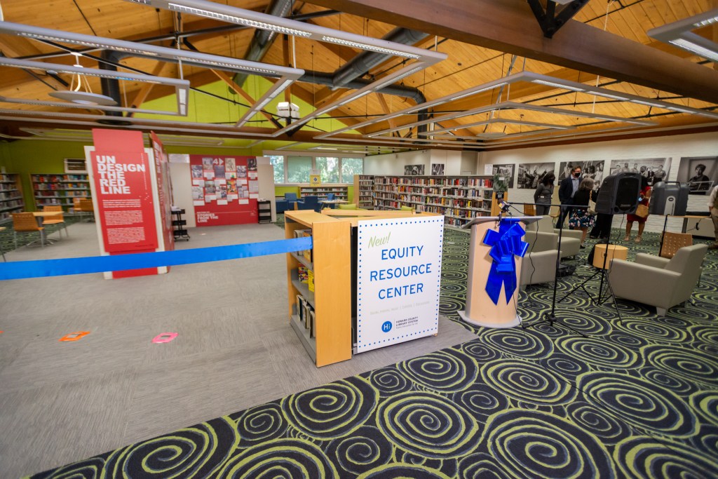 The Equity Resource Center at the Miller Branch, with a sign and podium for the ribbon cutting ceremony and bookshelves and the "Undesign the Red Line" exhibit in the background.