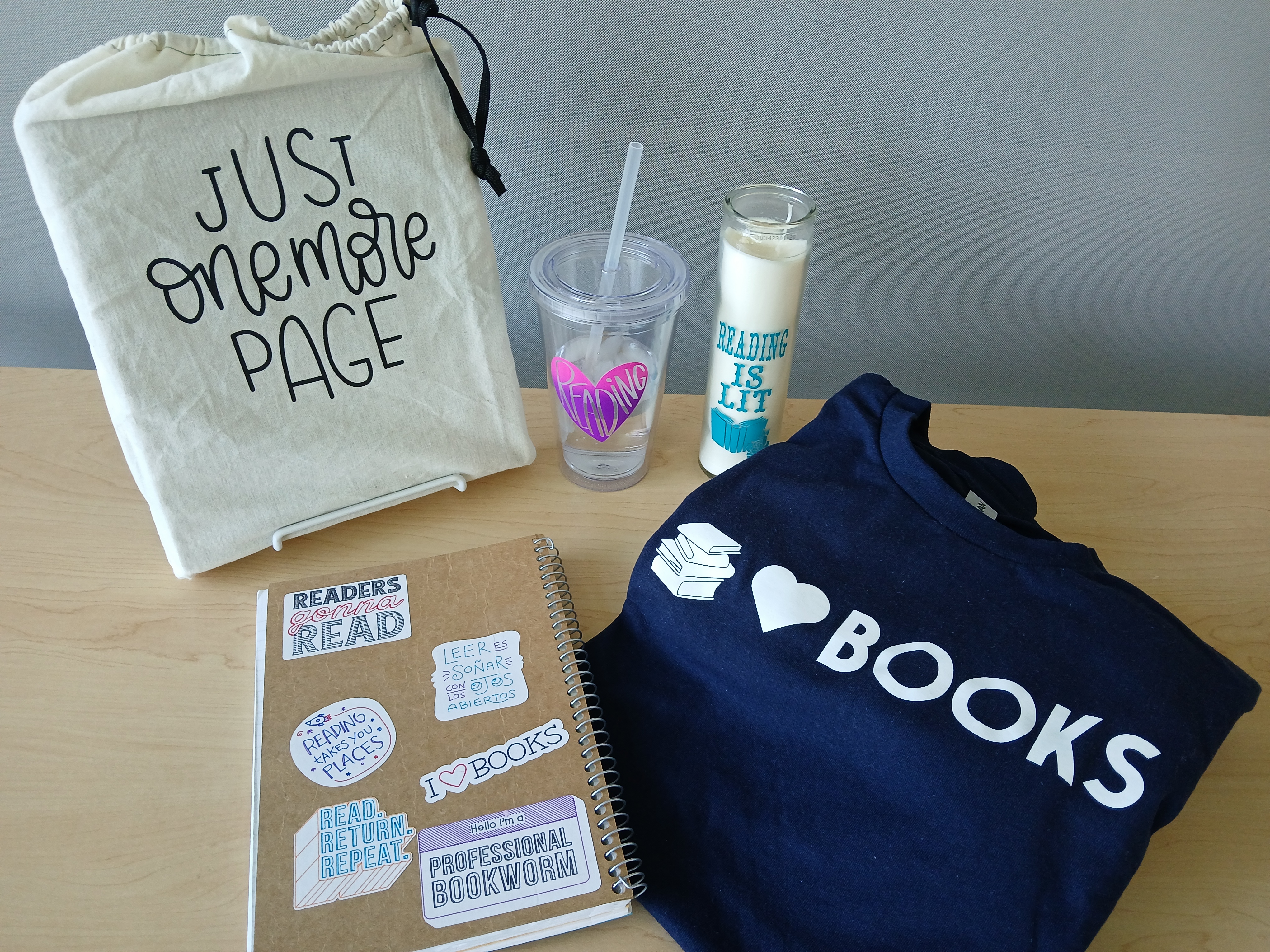 A variety of crafted items promoting reading: a canvas bag, a reuable water cup with straw, a tall candle in glass, a navy blue Tshirt, and a