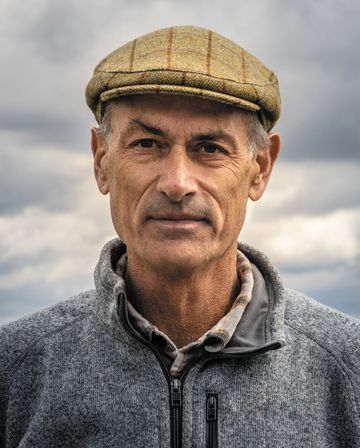 The author is an older White man wearing a tweed driving cap and medium grey zip up jacket, with grey clouds behind him.