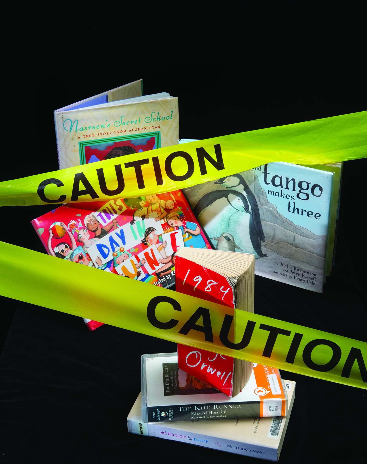Yellow caution tape stretches across an arrangement of books, all of which are perennially on the challenged book list. They include 1984 by George Orwell, The Kite Runner by Khaled Hosseini, and Tango Makes Three.