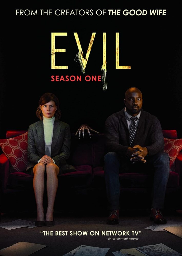 The DVD cover depicts a man and a woman seated side by side on an orange couch with cushions, wearing professional dress; a skeletal hand is reaching up between them from behind the sofa. The background is dark and there are papers scattered on the floor in front of them.