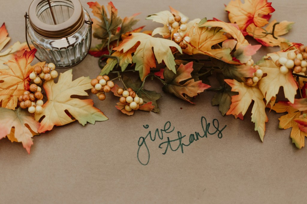 The photograph shows a table with an arrangement of autumn leaves and berries in shades of yellow, orange, green, and brown, with a votive candle in a glass and metal lantern-shaped holder and the words 'give thanks' written beneath them on the table in green cursive script.