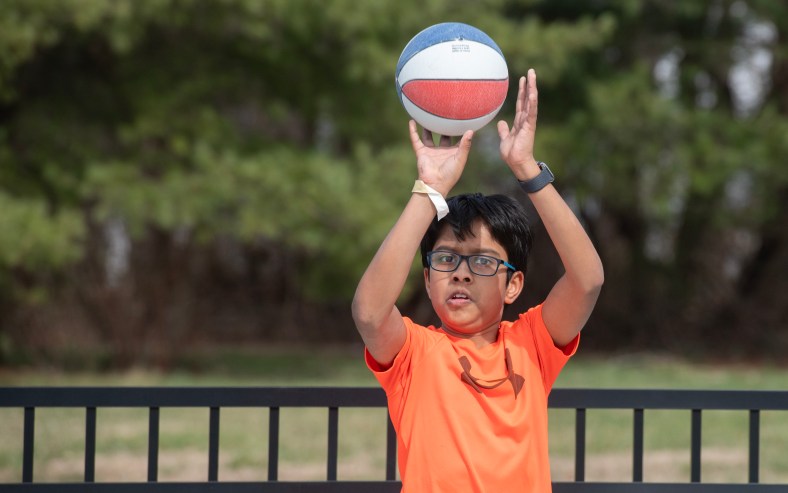 A boy with dark hair and glasses, wearing an orange T-shirt, gets ready to shoot a small basketball.