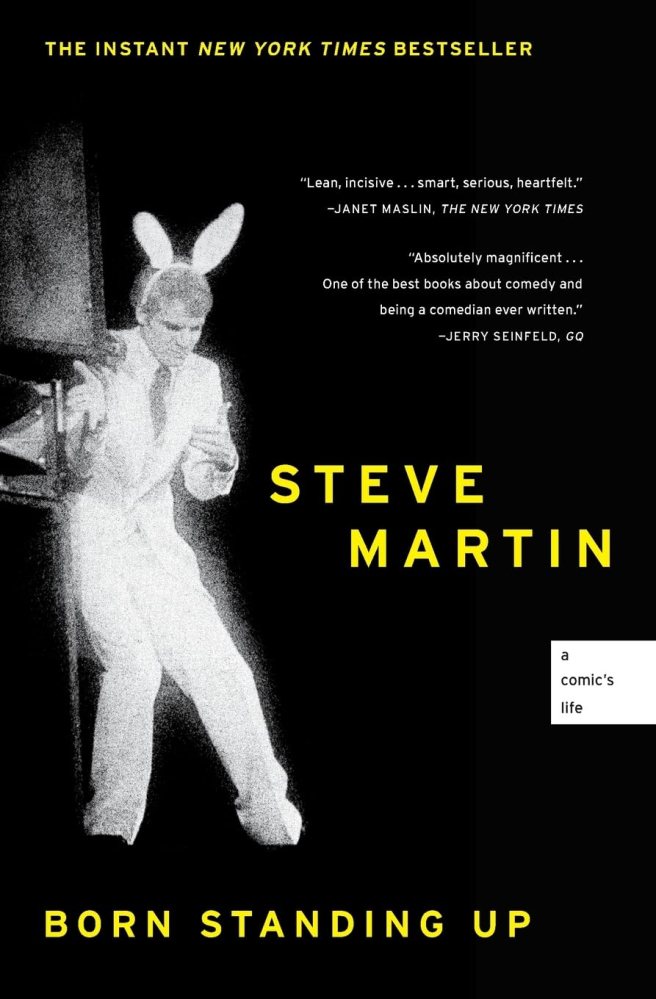 The photograph shows comedian and author Steve Martin wearing a white suit, a tie, and rabbit ears while on stage.