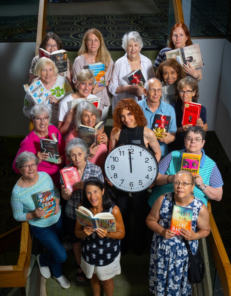 Members of the Noontime Bookclub stand on the steps at HCLS Central Branch, each holding a book and the group leader holding a clock set for noon.