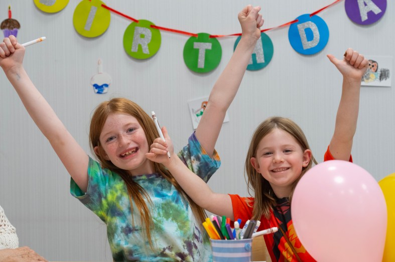 Two children celebrate at the Library's birthday party. They have their arms raised in front of decorations and balloons.