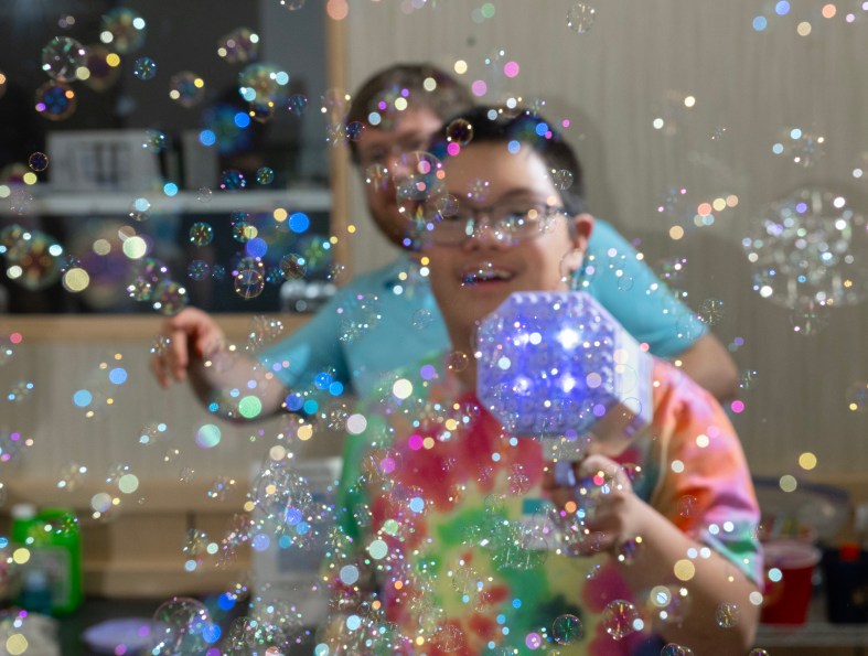 A young man with development disabilities smiles as he operates a bubble making machine. The air is full of small bubbles.